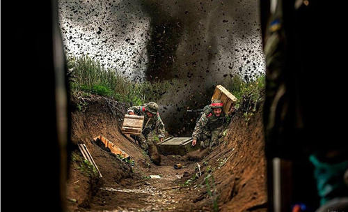 遭到炮击的乌军堑壕，两名士兵正冒着炮火搬运弹药箱。
